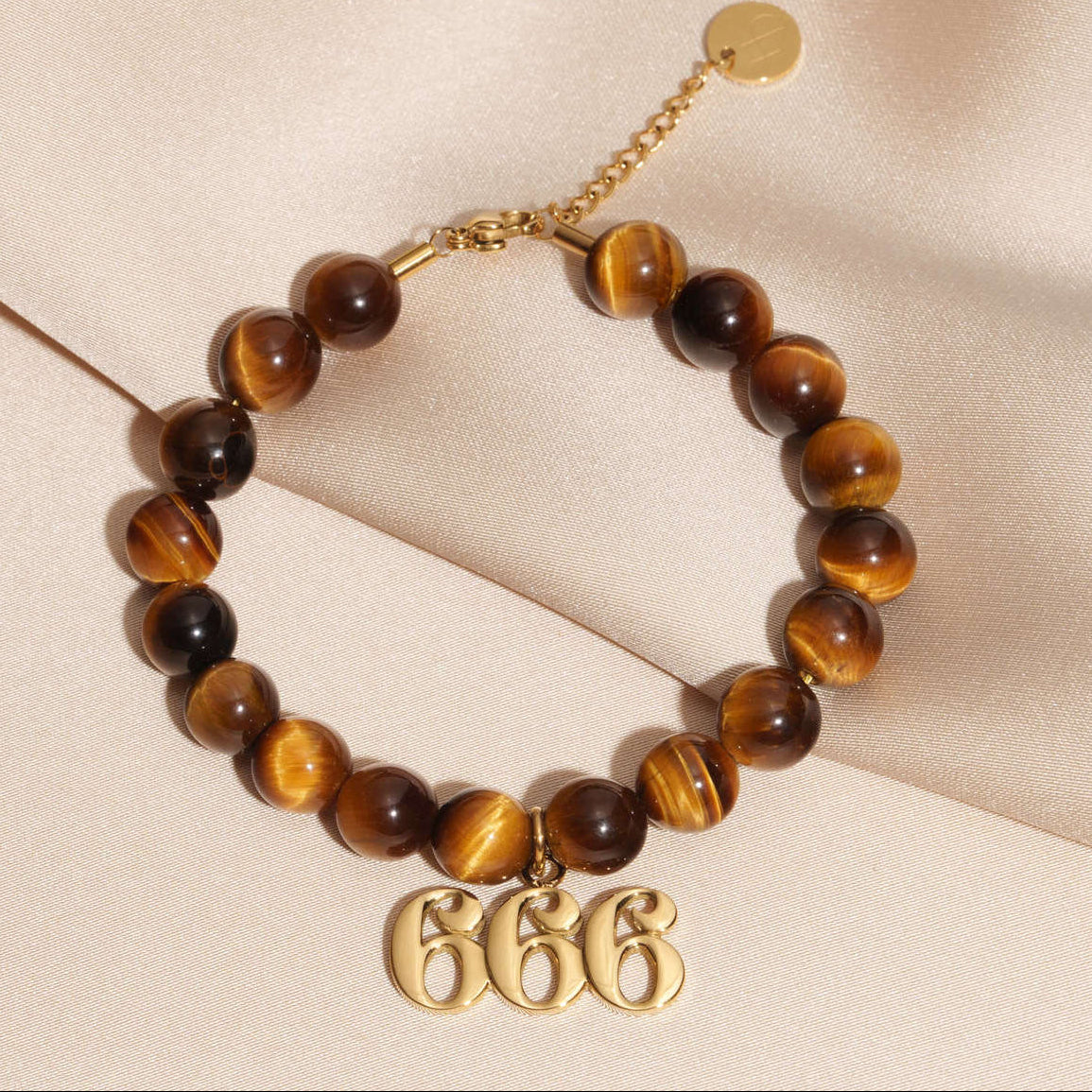 666 Gold charm on beaded Tigers Eye bracelet laying on silk
