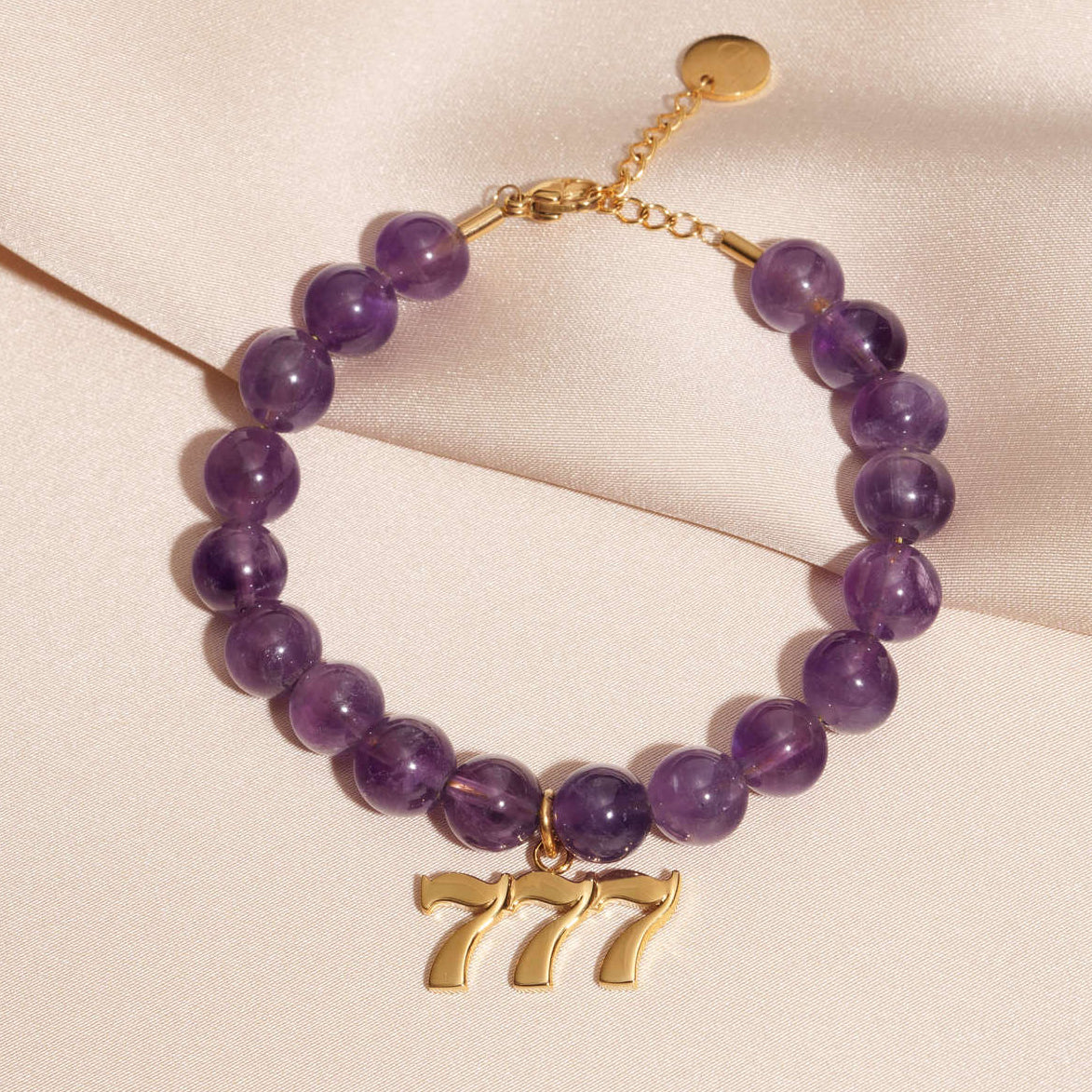 777 gold charm on beaded amethyst bracelet laying on silk
