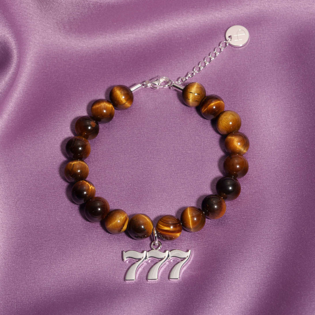 777 Silver charm on beaded Tigers Eye bracelet laying on silk
